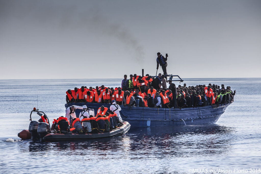 As some are taken to safety, other refugees and migrants on board a heavily overcrowded wooden boat eagerly wait for their chance to reach the safety of the Phoenix.