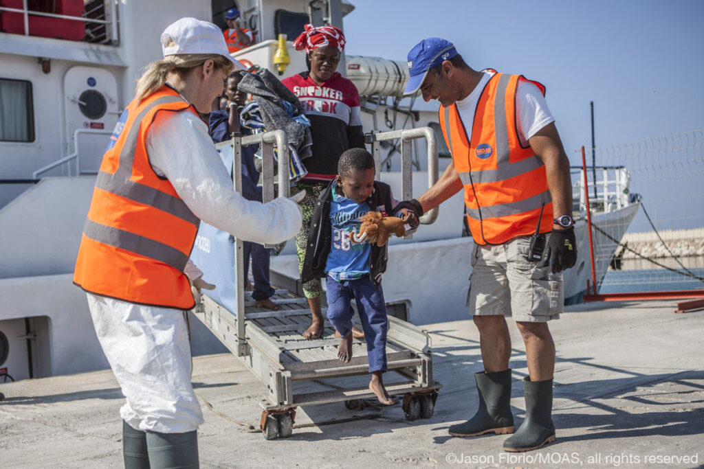 Crew member Mimmo, and MOAS co-founder and Director Regina Catrambone help a young boy and his mother off of the Phoenix as they disembark in Sicily.